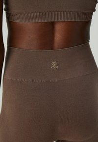 Close-up of midsection wearing ribbed brown seamless high-waist leggings and cropped top with small embroidered flower logo.