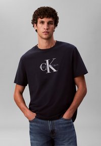 Tmavomodré bavlnené tričko s bielym logom Calvin Klein uprostred, s okrúhlym výstrihom a krátkymi rukávmi, v kombinácii s modrými rifľovými nohavicami.