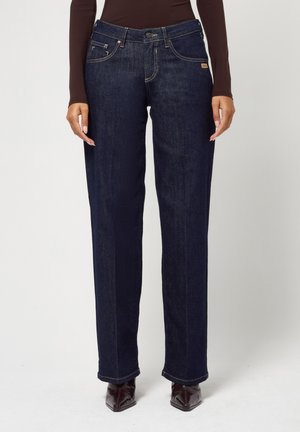 Person trägt dunkelblaue Jeans mit geradem Bein und braune spitze Schuhe, steht vor einem schlichten hellen Hintergrund.