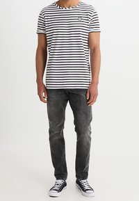 Camiseta a rayas blanco y negro con cuello redondo, mangas cortas y un logo circular en el pecho; combinada con pantalones vaqueros oscuros y zapatillas.