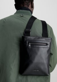 Borsa messenger in pelle nera con tasca frontale con zip, tracolla regolabile e dettaglio del logo; indossata sopra una giacca verde con una texture liscia.