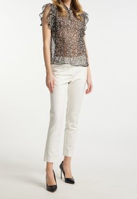 Blouse noire transparente avec imprimé floral, manches volantes et col, assortie à un pantalon blanc ajusté et des talons noirs.