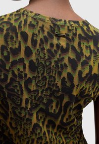 Närbild av en person som bär en grön, brun och svart leopardmönstrad topp med en liten metallmärke på den övre delen av ryggen.