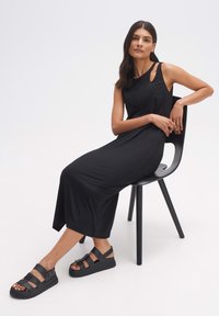 Zwarte, mouwloze maxi-jurk met uitsnijdingen op de schouders, gemaakt van een gladde stof. Gecombineerd met chunky zwarte sandalen op een stoel.