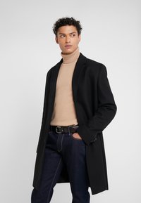 Cappotto di lana nero con una vestibilità sartoriale e rever a spillo, indossato su un dolcevita beige, abbinato a jeans in denim scuro e una cintura marrone.