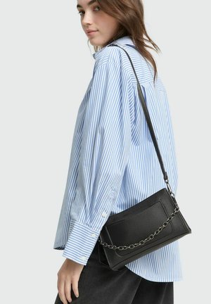 Schwarze Leder-Umhängetasche mit strukturierter Oberfläche, silberner Kettenriemen und kantigem Design, neben einem blau gestreiften Hemd präsentiert.