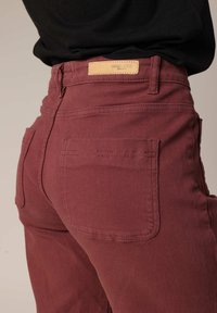 Jean bordeaux taille haute, avec poches arrière et une étiquette en cuir cousue sur la taille. Fabriqué en denim résistant.