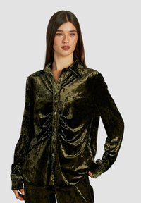 Camisa de terciopelo verde oscuro con cuello, mangas largas, cierre de botones en el frente y detalles fruncidos en el pecho. Textura suave.