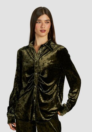Camicia in velluto verde scuro con colletto, maniche lunghe, chiusura a bottoni e dettagli arricciati sul petto. Texture liscia.