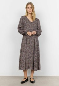 Lange V-hals jurk met pofmouwen, gemaakt van een patroon stof met paarse, roze en groene tinten. Gecombineerd met zwarte ballerina's.