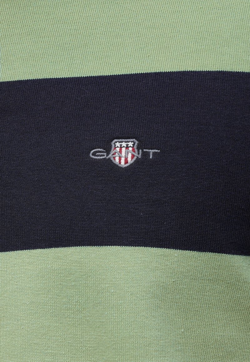 Tissu rayé vert et bleu marine avec le logo « GANT » et un petit écusson brodé rouge, blanc et bleu comportant trois étoiles blanches.