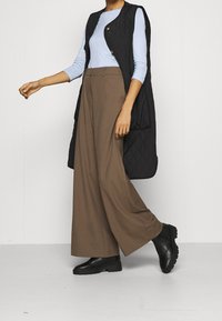 Persona che indossa un top a maniche lunghe azzurro chiaro, un gilet senza maniche trapuntato nero, pantaloni larghi marroni e scarpe nere chunky su uno sfondo semplice.