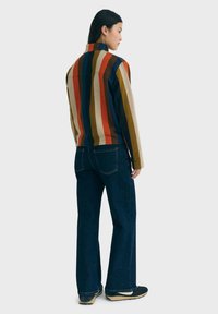 Giacca a righe multicolore con colletto alto, realizzata in tessuto morbido; abbinata a jeans a gamba larga blu scuro e scarpe nere.