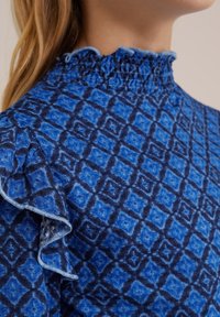 Blå mönstrad blus med smockad halsringning och volangärmar. Designen består av diamantformer i olika nyanser av blått.