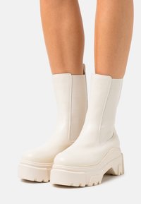 Jambes portant des bottes à plateforme blanches, mi-mollet, avec des semelles épaisses et des panneaux latéraux élastiques, présentées sur un fond clair et uni.