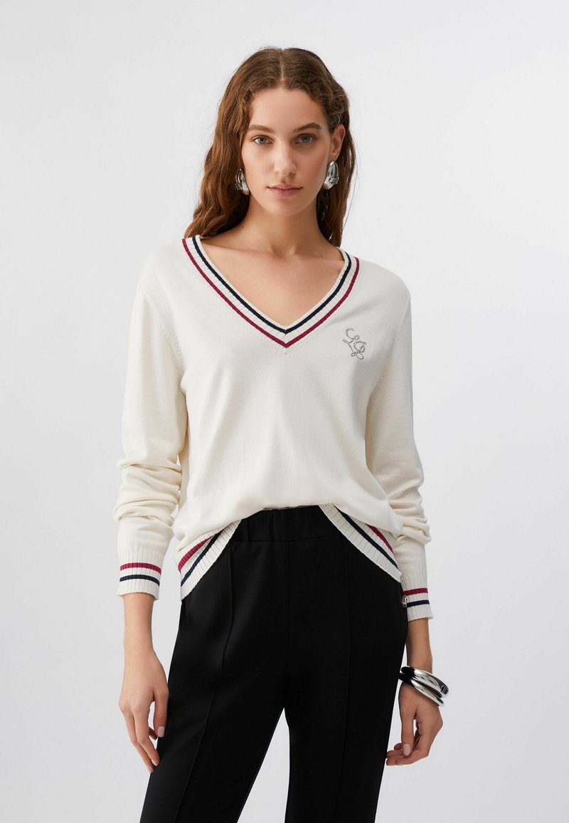 Femme aux longs cheveux bruns portant un pull blanc à col en V avec des rayures rouges et noires ainsi qu'un pantalon noir, accessoirisée de boucles d'oreilles et bracelets argentés.