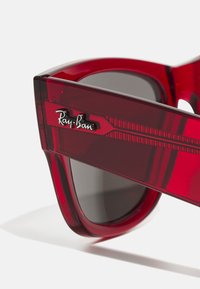 Ochelari de soare Ray-Ban roșii cu un cadru transparent lucios, lentile întunecate și detalii cu logo argintiu pe lateral, cu accente geometrice.