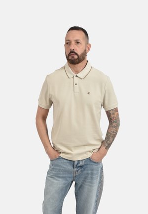 Hombre con tatuajes que lleva una camisa polo beige y vaqueros azules, de pie con las manos en los bolsillos, mirando hacia un lado con un fondo liso.