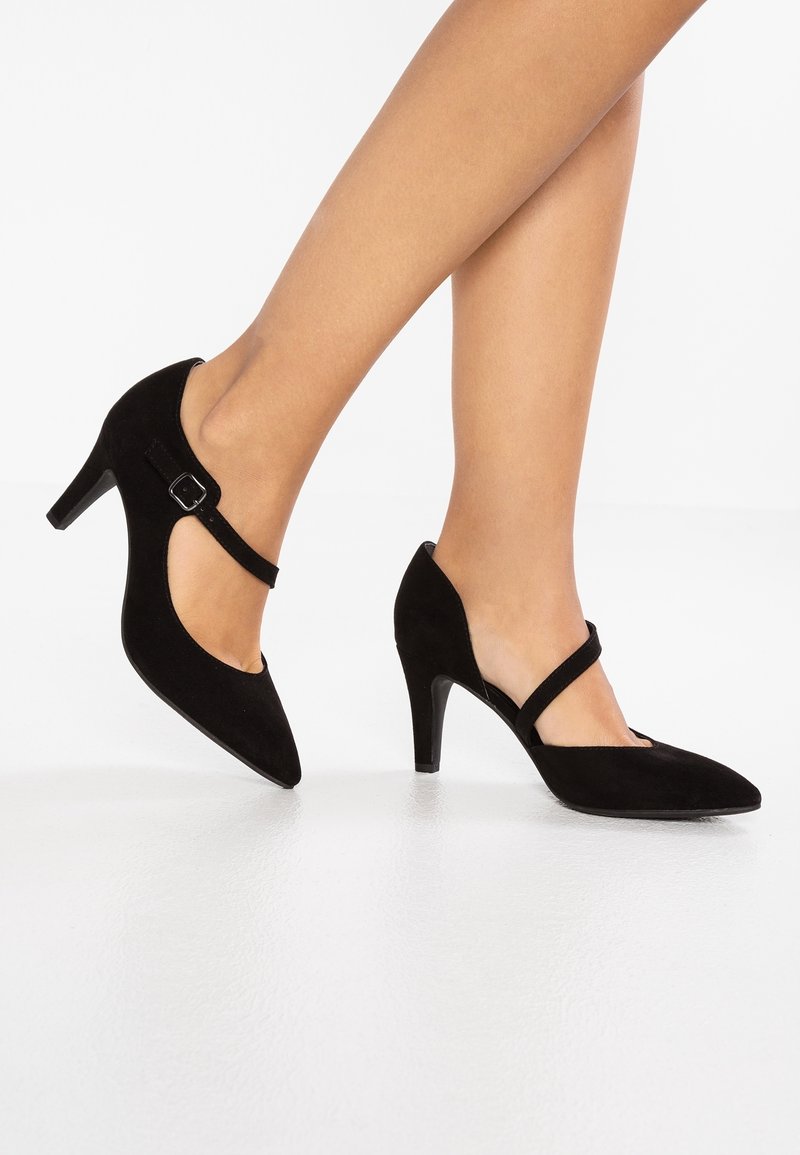Escarpins à talons hauts en daim noir avec bouts pointus et bride à la cheville. Présentent un talon fin et un design découpé sur les côtés pour plus de détails.