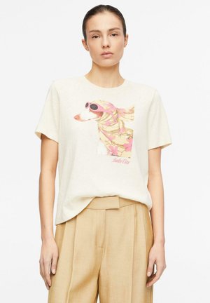 Kobieta ubrana w beżowe spodnie i biały t-shirt z grafiką psa noszącego różowe okulary przeciwsłoneczne i kwiatową chustę na głowie.