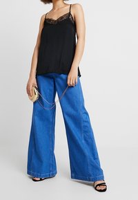 Camisole en soie noire avec bordure en dentelle, associée à un jean denim bleu à jambes larges et des sandales noires à talons. Accessoires compris : un petit sac à main rond.