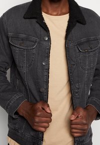 Svart denimjacka med fårskinnskrage, utrustad med två bröstfickor, metallknappar och synliga sömmar. Bärs över en beige skjorta.