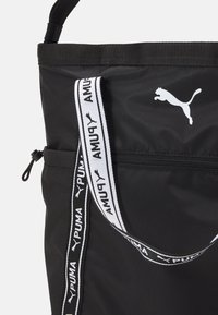Melnas auduma rokas soma ar baltu Puma logo, kurai ir melnbaltas uzšūtas siksnas un rāvējslēdzēja kabata priekšpusē.