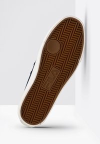 Suola di sneaker navy con una suola in gomma marrone strutturata, caratterizzata da un motivo a spina di pesce e un bordo laterale color crema con un logo.