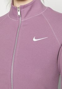 Lila gerippte Zip-Jacke mit hohem Kragen, versehen mit einem silbernen Reißverschluss und einem weißen Nike-Logo auf der linken Brustseite.