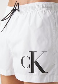 White swim shorts made of lightweight material, featuring an elastic waistband with a black drawstring and a large black "ck" logo on the side.