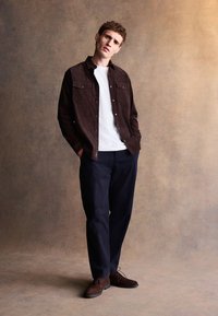 Brązowa koszula sztruksowa na białym t-shircie, granatowe spodnie i brązowe zamszowe buty. Model stoi z rękami w kieszeniach na tle z teksturą.