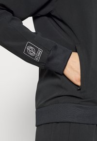 Chaqueta negra con un acabado suave, que presenta un bolsillo con cremallera y puños acanalados. Un parche con el logo en la manga añade un detalle de diseño.