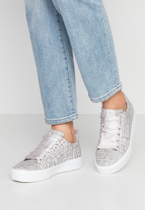 Personne portant un jean bleu clair et des baskets grises à motif crocodile avec de larges lacets en satin, se tenant sur un sol blanc uni.