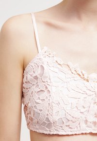 Kvinna som bär en blek rosa spets-brautlette med blommönster och smala axelremmar, som visar övre bröst och axel.