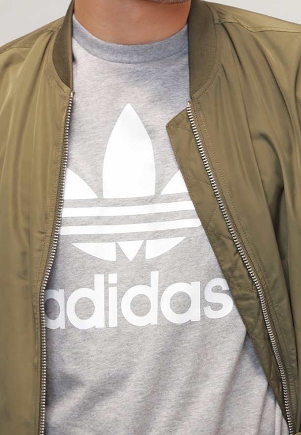 Vihreä oliivinvärinen bomber-takki vetoketjulla, harmaan t-paidan päällä, jossa on valkoinen Adidas-logo ja kolme vaakasuoraa raitaa.