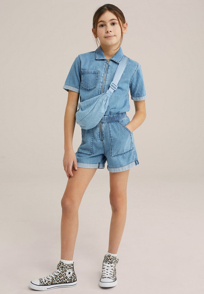 Jeansovski kombinezon v svetlo modri barvi s kratkimi rokavi, z zadrgo spredaj in elastičnim pasom. Združen z športnimi copati v leopardjem vzorcu.