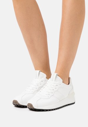 Legs wearing white lace-up sneakers with black tread soles on a white background, showing front and side views.