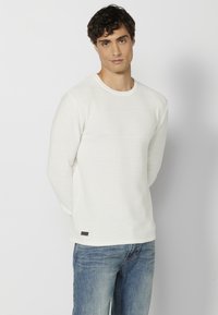 Witte lange mouwen shirt met een gestructureerd, gestreept patroon. Heeft een ronde hals en een klein logo-accent bij de zoom. Draagt met blauwe jeans.