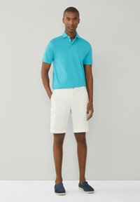 Man i turkos pikétröja, vita cargoshorts och mörkblå slip-in skor som står mot en enkel ljusgrå bakgrund.