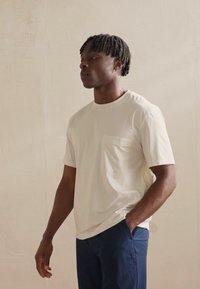 Svijetlosmeđi pamučni T-shirt s džepom na prsima, kratkim rukavima i okruglim izrezom, u kombinaciji s tamnoplavim hlačama. Glatka tekstura.