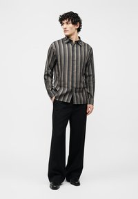 Νεαρός με σγουρά μαλλιά φορώντας μακρυμάνικο πουκάμισο με σχέδιο, μαύρο παντελόνι με φαρδιά πόδια και μαύρα παπούτσια, στέκεται με τα χέρια στις τσέπες.