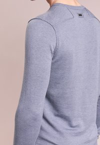 Homme portant un pull en tricot à manches longues de couleur violet clair, vu de dos et de côté sur un fond beige uni.