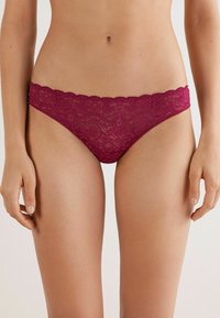 Perizoma in pizzo borgogna con bordi a scallop, caratterizzato da intricate fantasie floreali e una texture semi-trasparente. Progettato per comfort e stile.
