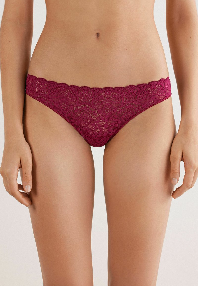 Perizoma in pizzo borgogna con bordi a scallop, caratterizzato da intricate fantasie floreali e una texture semi-trasparente. Progettato per comfort e stile.