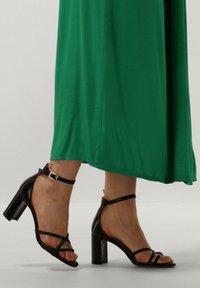 Zwarte haksandalen hebben een glanzende afwerking, een strappy ontwerp met enkelgesp, en een stevige blokhak, gecombineerd met een groene outfit.