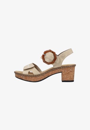 Beige sandal with a cork wedge heel, featuring an adjustable strap and a large brown plastic buckle accent. Soft upper material and open-toe design.