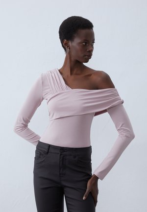 Rosa langermet topp med et asymmetrisk design, med drapert stoff på den ene skulderen. Kombinert med sorte tettsittende bukser. Glatt tekstur.