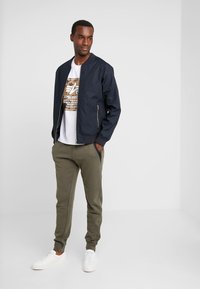 Marine bomberjack met ribbels aan de mouwen, wit graphic T-shirt, olijfgroene joggers en witte sneakers op een model voor een effen achtergrond.