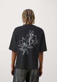 Person med blont, tvinnat hår bär en svart oversized t-shirt med stort vitt blomsterlinjeteckning på baksidan, ihop med blekta svarta jeans.