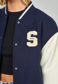 Marinblå ull varsity-jacka med krämfärgade läderärmar, med en krämfärgad "S"-lapp och ribbad krage med vita ränder. Framsnap stängning.